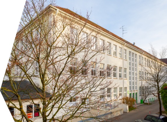 Das Bild zeigt ein mehrstöckiges weißes Gebäude mit zahlreichen Fenstern. Das Gebäude ist von blattlosen Bäumen umgeben, was auf Winter oder frühen Frühling hinweist.